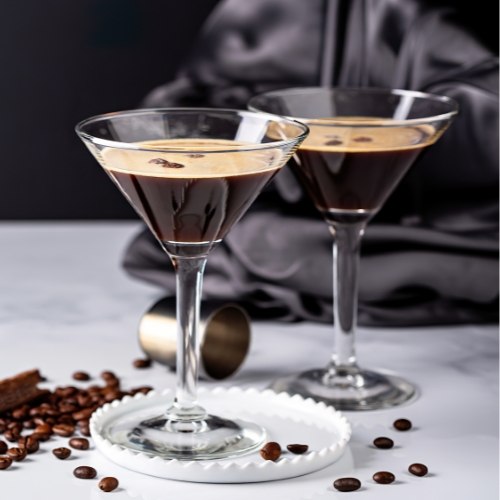 Deux verres à martini contenant un Espresso Martini , avec une mousse onctueuse et des grains de café en garniture, posés sur un plateau blanc avec des grains de café en fond 
