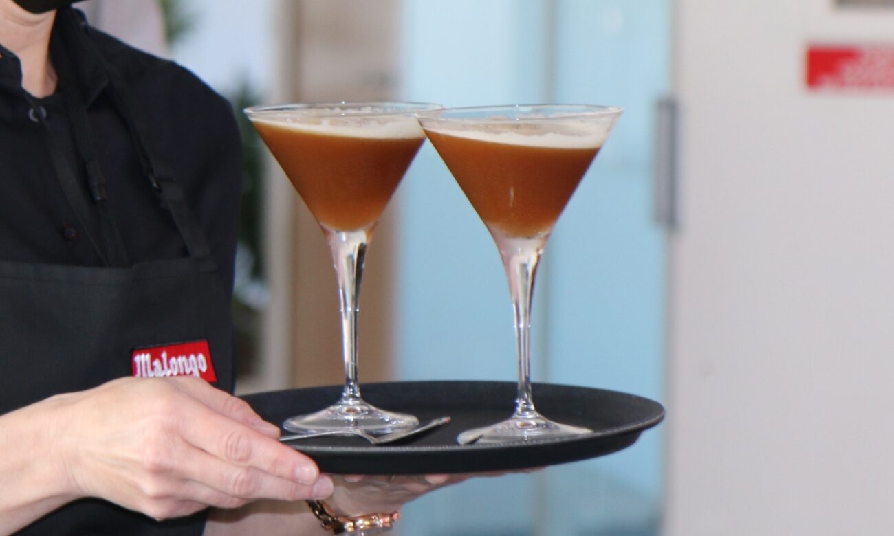 Plateau de barista Malongo présentant deux verres à cocktail espresso martini - boisson froide à base de café en grain torréfié artisanalement, crème onctueuse et bulles visibles - servi par un professionnel en tenue noire avec tablier marqué du logo Malong
