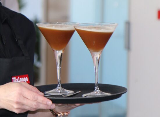 Plateau de barista Malongo présentant deux verres à cocktail espresso martini - boisson froide à base de café en grain torréfié artisanalement, crème onctueuse et bulles visibles - servi par un professionnel en tenue noire avec tablier marqué du logo Malong