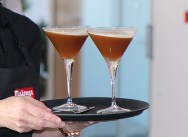Plateau de barista Malongo présentant deux verres à cocktail espresso martini - boisson froide à base de café en grain torréfié artisanalement, crème onctueuse et bulles visibles - servi par un professionnel en tenue noire avec tablier marqué du logo Malong
